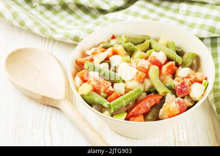 Un mélange de légumes surgelés assortis dans un bol et avec une cuillère en bois sur une table en bois blanc Banque D'Images