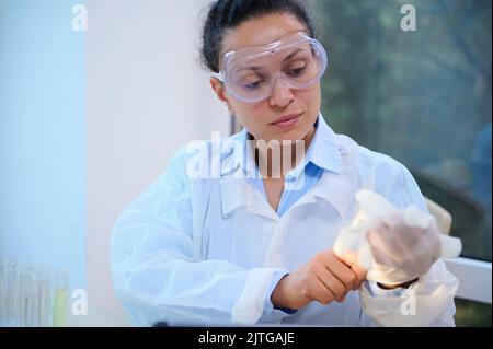 Une scientifique féminine, employée de laboratoire médical en blouse blanche et lunettes de protection met des gants de sécurité chirurgicaux Banque D'Images