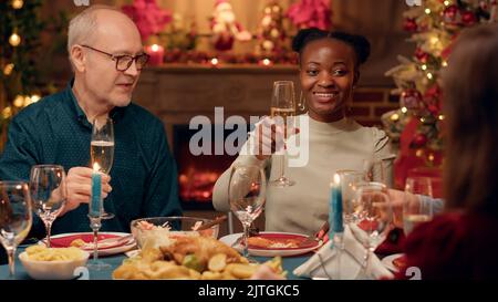 Riant avec enthousiasme, les gens se réjouissant de prendre des verres de champagne tout en appréciant le dîner de Noël. Membres de famille multiethniques festifs célébrant ensemble les vacances d'hiver tout en buvant du vin mousseux. Banque D'Images