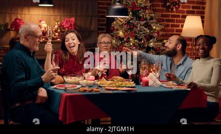 Souriant, les gens rient avec cœur lorsqu'ils parlent à l'appareil photo lors du dîner de Noël. Des membres de la famille joyeux célèbrent ensemble les vacances d'hiver tout en appréciant le temps ensemble à la maison. Cliché sur trépied Banque D'Images