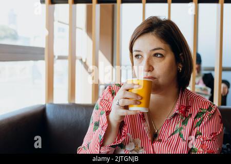 jeune femme latine de race blanche argentine aux cheveux courts et aux vêtements décontractés, elle boit du café et pense, assis dans un restaurant de restauration rapide Banque D'Images