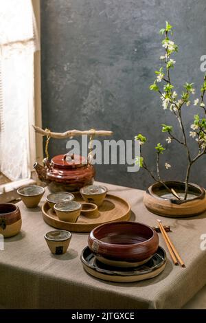 Table de style japonais asiatique avec vaisselle en céramique artisanale vide, bols bruns et tasses sur la nappe en lin, décorée par ikebana Spring bloo Banque D'Images