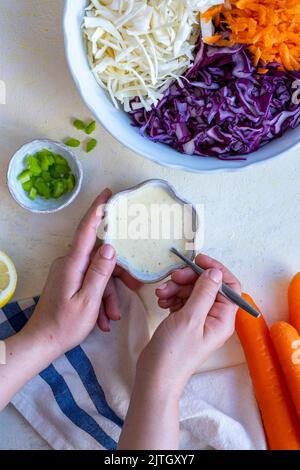 Femme mélange les mains de la sauce au yogourt aux coleslaw, un bol de chou rouge en tranches, de chou blanc et de carotte râpée sur le côté. Banque D'Images