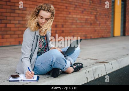 Portrait d'un étudiant ciblé avec des cheveux bouclés faisant ses devoirs, écrivant dans un ordinateur portable, assis avec les jambes croisées à l'extérieur. Été, Internet Banque D'Images