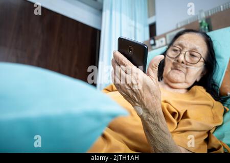 Femme d'Asie du Sud-est, patiente sénichée sur le lit d'hôpital et se servant d'un smartphone pour se divertir Banque D'Images