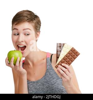 Changez d'avis, changez de corps. Photo studio d'une jeune femme en forme qui décide de manger du chocolat ou une pomme sur fond blanc. Banque D'Images