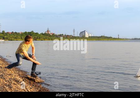 Jeune homme en T-shirt jaune et jeans sur la plage de pierre, sautant des pierres sur la rivière. Beau homme aux rayons du soleil sur les pierres chaudes et claires de la soirée d'été sautant Banque D'Images