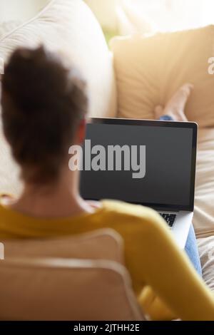 Voir le monde depuis le confort de son canapé. Vue arrière d'une jeune femme utilisant son ordinateur portable sur le canapé à la maison. Banque D'Images