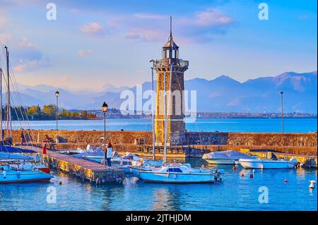 Phare en pierre historique sur Diga Foranea brise-lames avec yachts et bateaux dans le port et les préalps de Garda dans le fond au coucher du soleil brumeux, Desenzano Banque D'Images