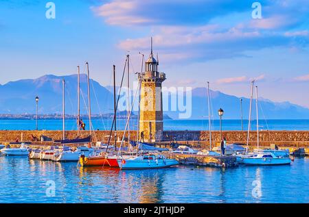 La soirée brumeuse sur le lac de Garde avec des yachts, le phare de Faro et les silhouettes de Garda Prealps en arrière-plan, Desenzano del Garda, Lombardie, Italie Banque D'Images