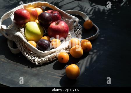 Fruits frais mûrs et baies sur fond de bois noir dans un sac en maille écologique. Style de vie. Ombres dures.lumière du soleil.récolte mûre de pommes. Cerises, abricots et prunes. Copier l'espace. Banque D'Images