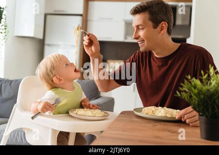 Bonne journée des pères. Vue latérale d'un papa qui donne un repas à bébé dans un bavoir vert avec spaghetti pendant le déjeuner à la table de cuisine, un enfant assis dans une fourchette de maintien pour chaise haute. Bonne paternité, premier repas, auto-alimentation Banque D'Images