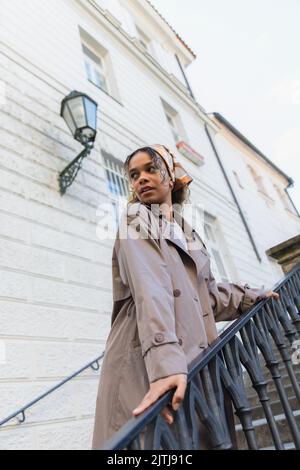 femme afro-américaine en foulard et en trench-coat branché touchant la main courante en se tenant sur les escaliers de prague Banque D'Images