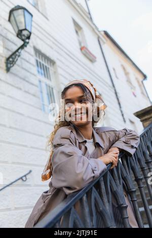 femme afro-américaine gaie en foulard et en trench-coat tendance touchant la main courante en se tenant sur les escaliers de prague Banque D'Images