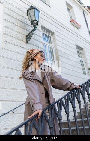 femme afro-américaine en foulard et trench-coat tendance touchant la main courante en se tenant sur les escaliers de prague Banque D'Images