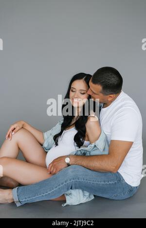 Jeune famille heureuse mari et femme attendent un bébé. Un homme embrasse une femme enceinte sur fond gris Banque D'Images