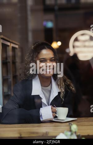 bonne femme afro-américaine tenant une tasse de café et souriant derrière le verre de fenêtre dans le café Banque D'Images
