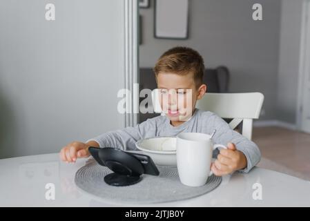 Un enfant, un garçon de cinq ans, prend le petit déjeuner à la table de cuisine et regarde des dessins animés sur son smartphone Banque D'Images