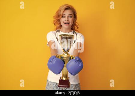 Femme joyeuse célébrant la victoire. Une femme en gants de boxe tient le trophée de la coupe gagnante du champion. Boxeur féminin primé isolé sur fond jaune. Banque D'Images