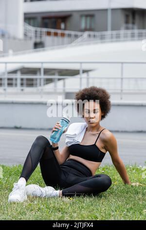 femme afro-américaine élancée dans des vêtements de sport noirs, assise sur la pelouse avec une serviette et une bouteille de sport Banque D'Images