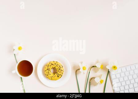 Le lieu de travail d'un blogueur ou d'un freelance à plat est une femme avec un clavier, une tasse de thé, des beignets sur une assiette et des fleurs de printemps jonquilles sur un dos léger Banque D'Images