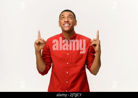 Concept publicitaire. Un gars afro-américain excité en chemise rouge pointe les doigts isolés sur le blanc. Souriant homme multiracial en prêtant votre attention ci-dessus à l'espace de copie Banque D'Images