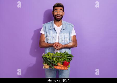 Photo d'un joli gars habillé gilet en denim tenant vert panier légumes vide espace isolé couleur violet fond Banque D'Images