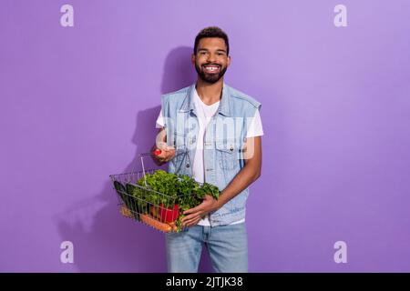 Photo de drôle mignon gars habillé gilet en denim tenant des légumes frais panier isolé couleur violet arrière-plan Banque D'Images