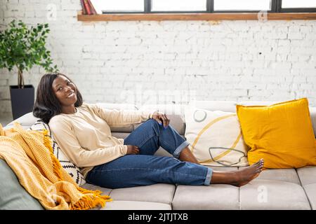 Femme en bonne santé assise sur le canapé confortable pur dans un appartement moderne, vêtue de vêtements décontractés et de gaies souriantes Banque D'Images