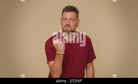 Agressif et enragé jeune homme en t-shirt rouge essayant de se battre à la caméra, en secouant le poing, la boxe avec l'expression, la punition. Adulte homme élégant gars isolé seul sur fond beige studio à l'intérieur Banque D'Images