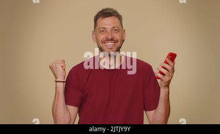 Joyeux joyeux joyeux jeune homme utiliser smartphone dactylographiant navigation dire wow oui, a découvert de grandes nouvelles de gain faisant le geste du gagnant. Homme de style adulte isolé sur fond de mur beige studio à l'intérieur Banque D'Images