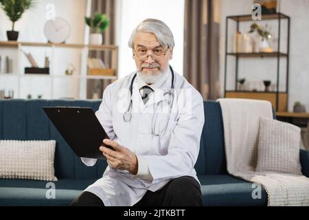 Médecin aux cheveux gris confiant en lunettes et blouse de laboratoire écrivant des notes sur le tableau à feuilles mobiles tout en posant à l'hôpital. Travailleur médical caucasien avec stéthoscope sur le cou debout à l'intérieur. Banque D'Images
