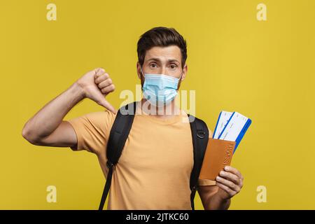 Mauvais service de l'agence de voyage. Homme contrarié tenant un passeport avec des billets, montrant les pouces vers le bas, portant un sac à dos et un masque facial Banque D'Images