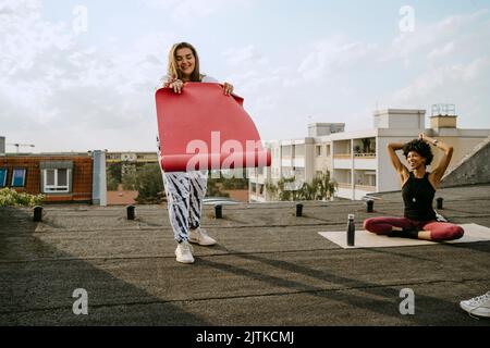 Une femme souriante pose un tapis d'exercice par une amie pratiquant le yoga sur le toit Banque D'Images