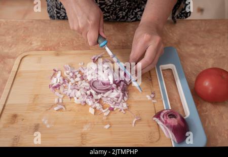 Vue de dessus des mains d'une femme hacher des oignons avec un couteau bleu sur une planche en bois au comptoir de la cuisine Banque D'Images