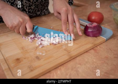 Gros plan sur les mains d'une femme hacher des oignons avec un couteau bleu sur une planche en bois au comptoir de cuisine Banque D'Images