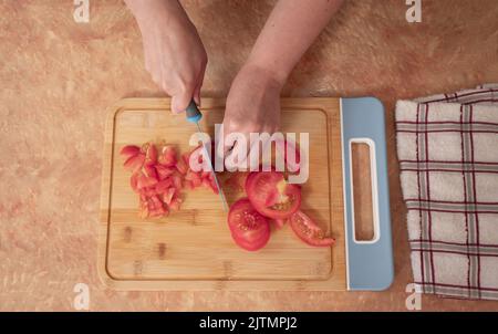 Vue de dessus des mains de femme hacher des tranches de tomate avec un couteau bleu sur une planche en bois au comptoir de cuisine Banque D'Images