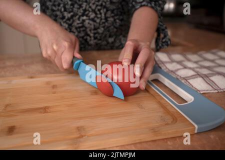 Gros plan des mains d'une femme coupant une tomate en tranches avec un couteau bleu sur une planche en bois au comptoir de la cuisine Banque D'Images
