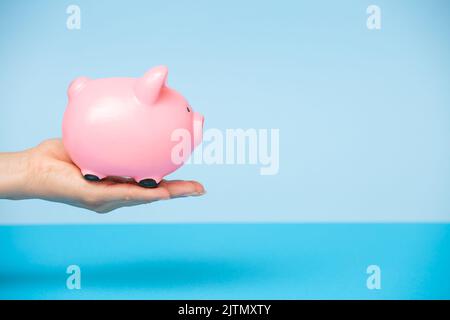Une main de femme tient une banque de porc rose sur un fond bleu.le concept d'investissement dans l'avenir, l'épargne, la planification financière.Copy espace Banque D'Images