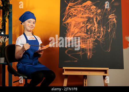 Portrait d'une petite fille qui peint des œuvres d'art sur toile, à l'aide d'un plateau colormix et d'une palette de peinture colorée. Artiste créant chef-d'œuvre avec couleur orange aquarelle, ayant des compétences et une vision artistique. Banque D'Images