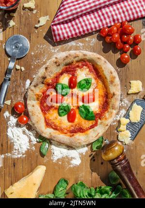Pizza napolitaine avec tomate et basilic. Servi sur une table en bois et entouré de tous les ingrédients. Scène de préparation. Parmesan et poivre. Banque D'Images