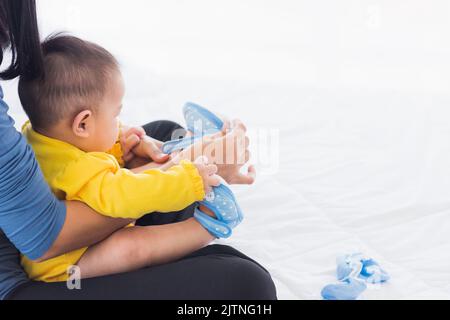 Une jeune mère asiatique assise sur le lit portant des chaussures à son bébé adorable petite fille tout-petit dans la chambre, maman aidant le petit bébé porte des chaussures Banque D'Images