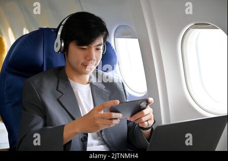 Beau jeune homme d'affaires asiatique ou passager masculin portant un casque sans fil et jouant à un jeu mobile pendant le vol. Banque D'Images