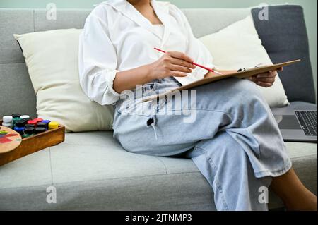 Décontractée asiatique femme assis sur le canapé dans son salon de maison, dessin et peinture de couleur de l'eau, faisant son beau travail d'art. Concept de passe-temps créatif Banque D'Images