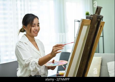 Belle et charmante femme asiatique millénaire tenant une palette, utilisant la peinture de pinceau sur le grand chevalet de toile dans son studio à la maison. Banque D'Images