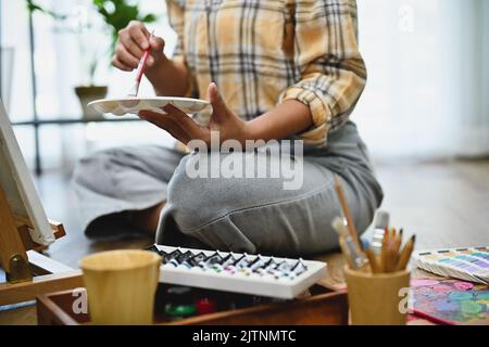 Artiste féminine talentueuse tenant une palette, dessinant et peignant son œuvre sur un chevalet de toile vierge dans son studio d'art. Image rognée et gros plan Banque D'Images