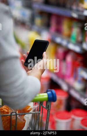 La commodité d'une liste d'achats numérique. Gros plan d'une femme qui vérifie sa liste d'achats numériques sur son téléphone portable dans une épicerie. Banque D'Images