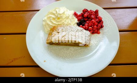 Strudel aux baies maison en tranches, servi avec des raisins de Corinthe rouges frais et du sucre en poudre sur une assiette de crème fouettée Banque D'Images