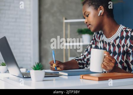 Jeune femme d'affaires afro-américaine prospère et attirante assise à table de bureau, en train de résoudre des problèmes mathématiques, avec des manuels scolaires et un appareil électronique Banque D'Images