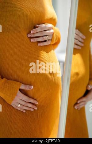 Femme enceinte en robe regarde son reflet dans le miroir. Concept de l'amour et des soins pendant la grossesse, l'attente et le bébé né. Banque D'Images